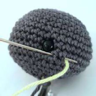 Needle coming out two stitches behind the safety eye of a crochet bat head, with lime yarn