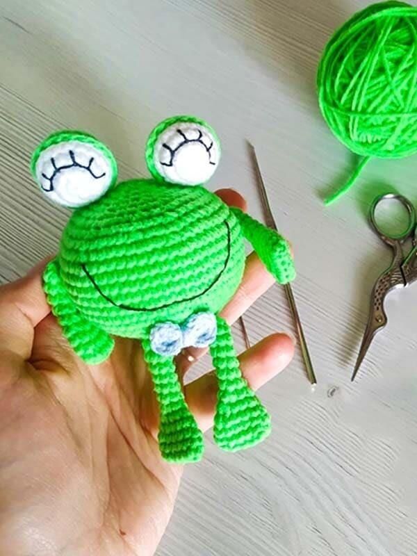 Green crochet frog with white raised eyes, long legs and a light blue bow, held in a hand