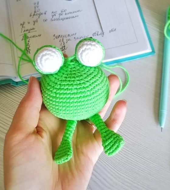 Round green crochet frog body with both long legs joined at the bottom and the raised eyes pinned on top