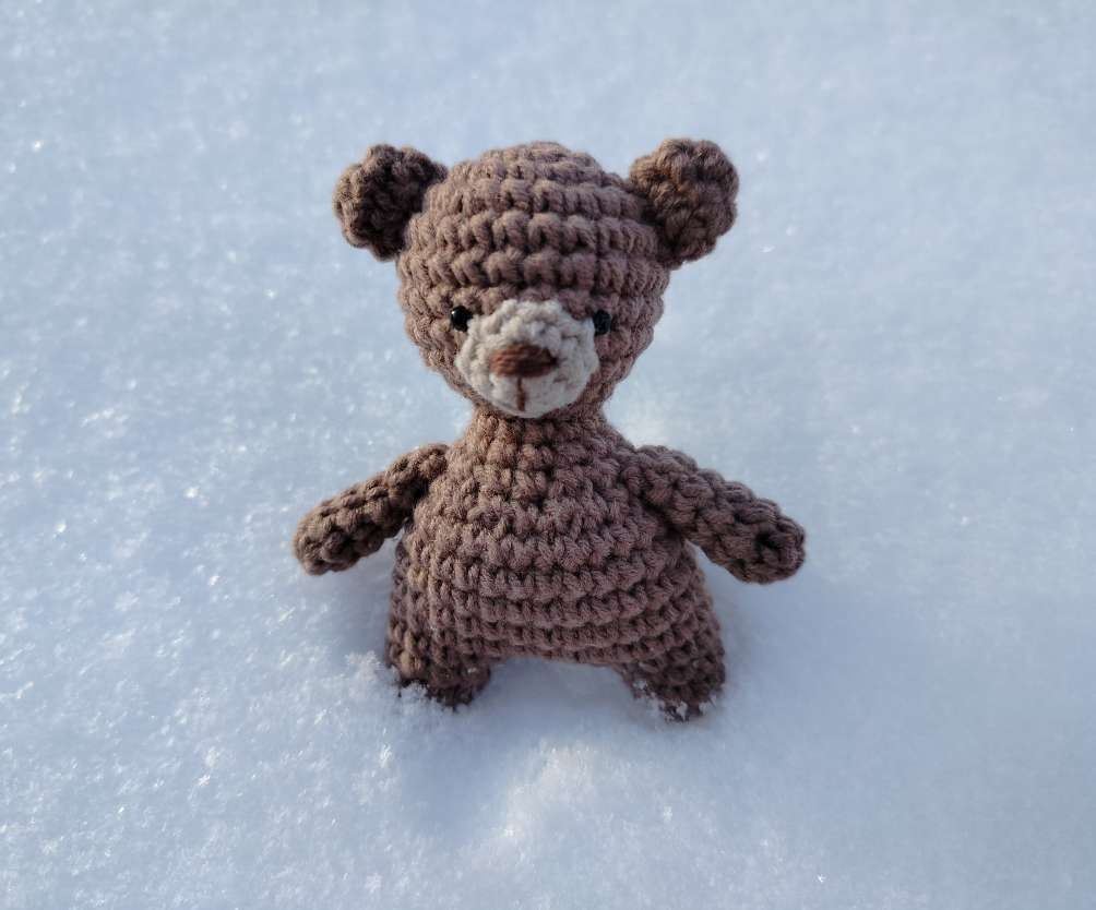 Small brown crochet teddy bear with round ears and a light muzzle, sitting on snow
