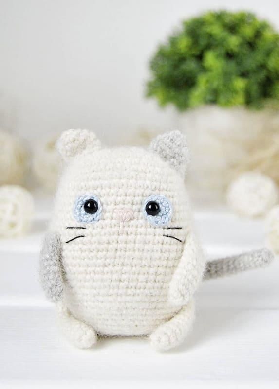 Small cream crochet cat with one grey ear, one grey arm, a grey tail, blue eye irises and embroidered whiskers