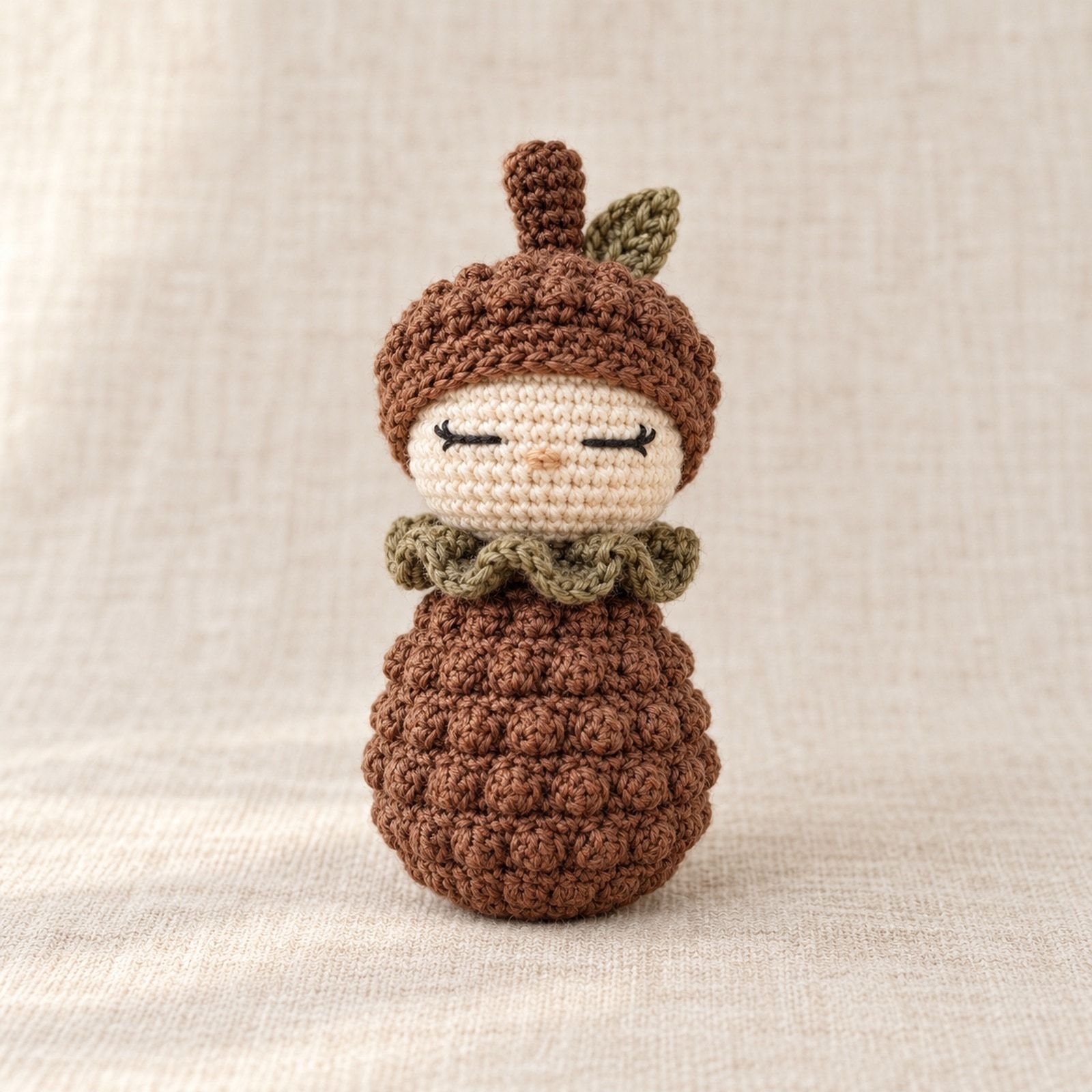 Woodland crochet figure with a cream head, brown bobble body and cap, an olive leaf and ruffled collar.