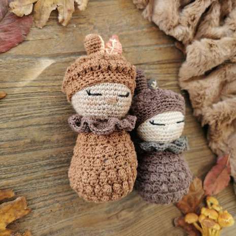 A brown and a pink crochet pine cone baby doll side by side on a wooden table with autumn leaves
